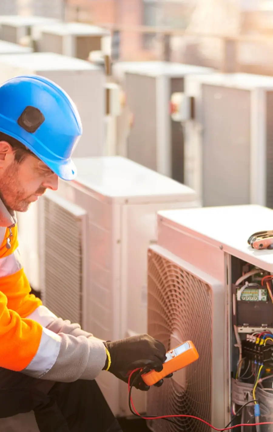 A technician in a hard hat testing an outdoor air-conditioning unit with a multimeter